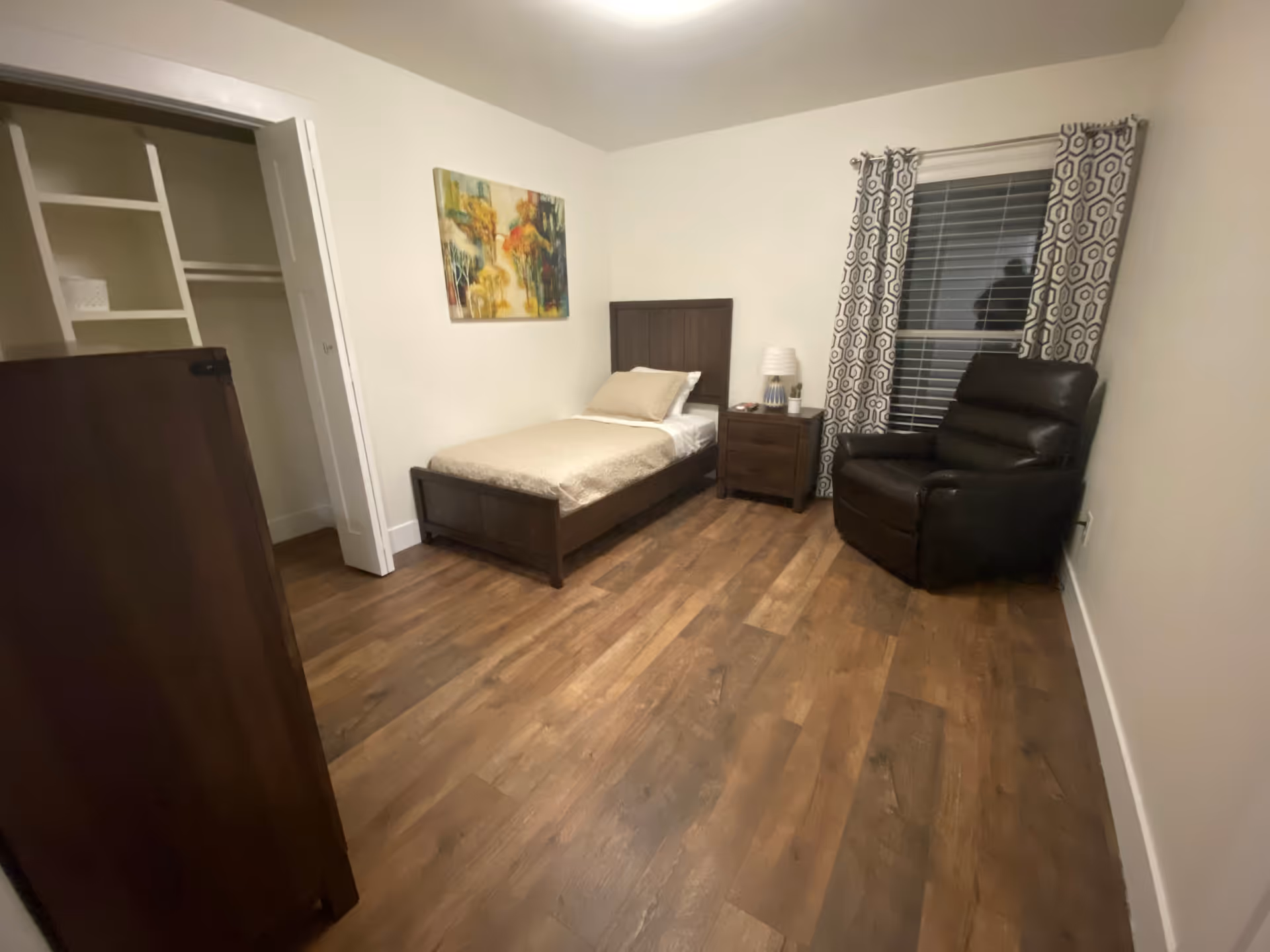 A simple bedroom with a single bed against the wall, a nightstand with a lamp, a dark brown recliner chair near a window with patterned curtains, a wooden dresser, and an open closet with shelves. The floor is wooden and the walls are painted white with a colorful abstract painting hanging above the bed.