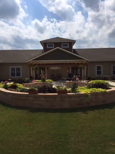 Outdoor view of a single-story building with a covered porch area, surrounded by a circular raised garden bed filled with various plants and flowers, under a partly cloudy sky.