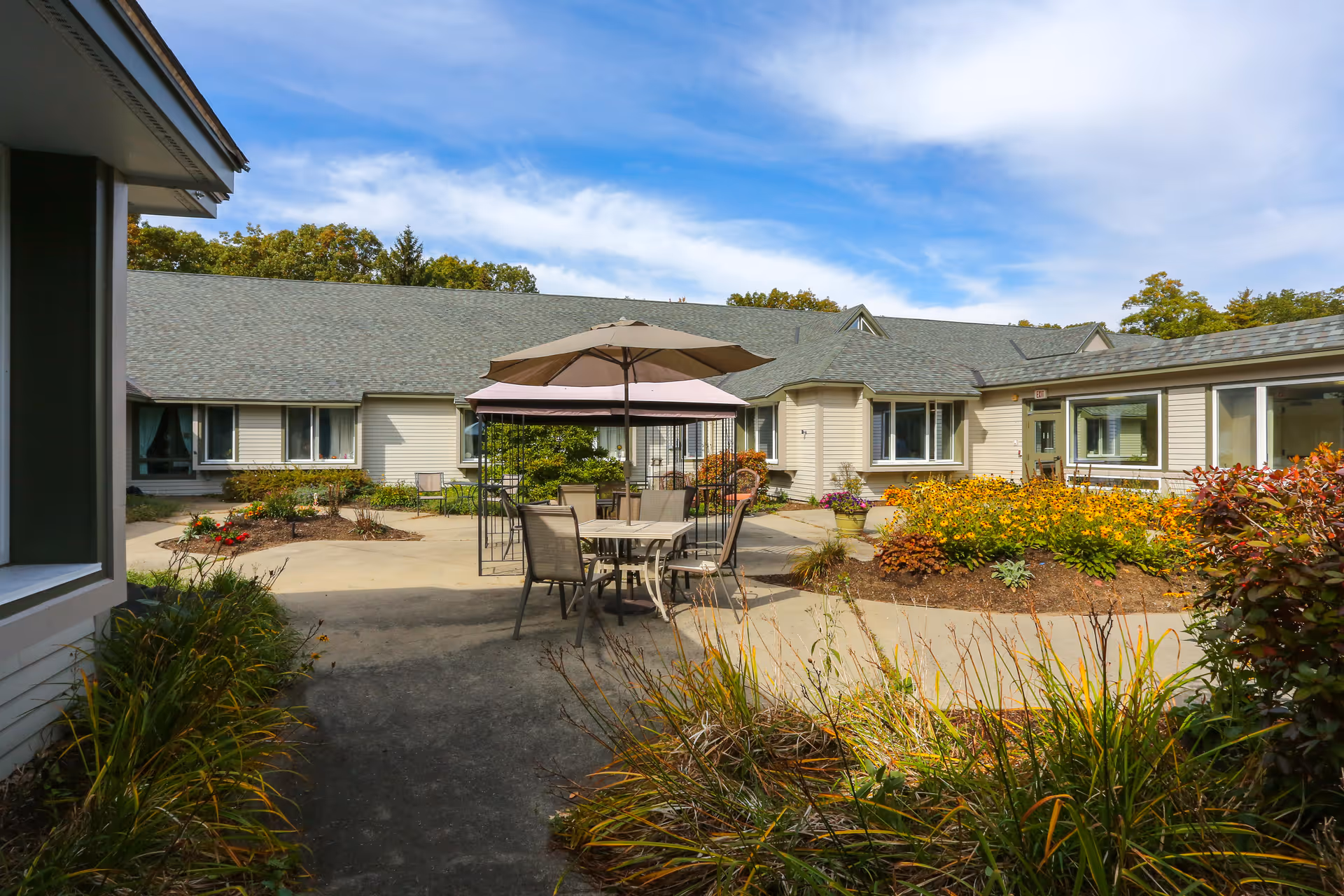 Outdoor courtyard area of a senior living facility with a round table and chairs under a large umbrella, surrounded by flower beds and shrubs, with a single-story building and blue sky in the background.