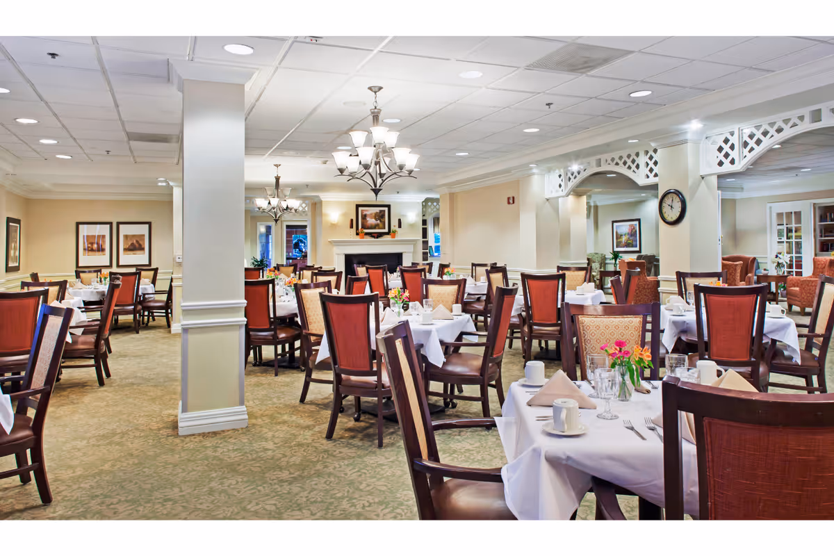 A spacious dining room with multiple tables covered in white tablecloths, set with napkins, cups, glasses, and silverware. The room features wooden chairs with red and patterned upholstery, chandeliers hanging from the ceiling, framed artwork on the walls, and a fireplace in the background. The carpet is green with a subtle pattern, and there are decorative white lattice arches along one wall.