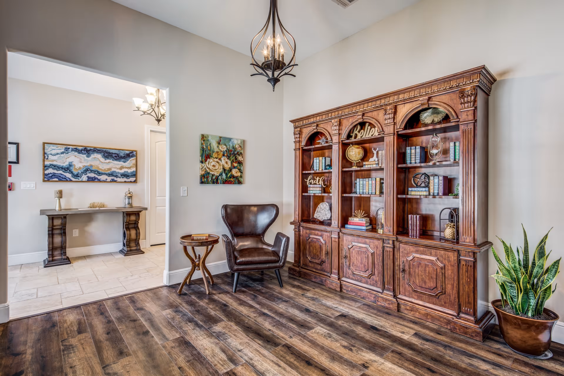 A cozy interior room with wooden flooring featuring a large ornate wooden bookshelf filled with books and decorative items. A dark leather armchair and a small round wooden side table are placed near the bookshelf. The walls are light-colored with two paintings hanging, and there is a chandelier hanging from the ceiling. A potted plant is positioned on the floor to the right of the bookshelf. An adjacent room with a console table and artwork is visible through an open doorway.