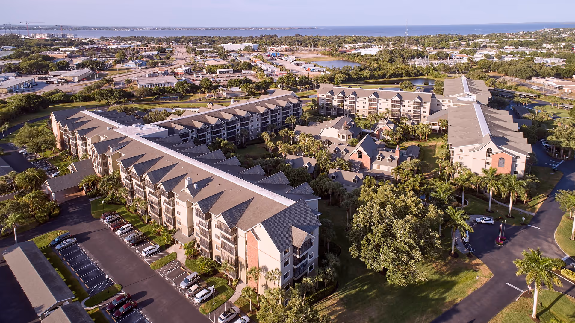 Aerial view of South Port Square senior living facility showing multiple large residential buildings surrounded by trees, parking lots, and roads. The facility is located near a body of water with a suburban area in the background.