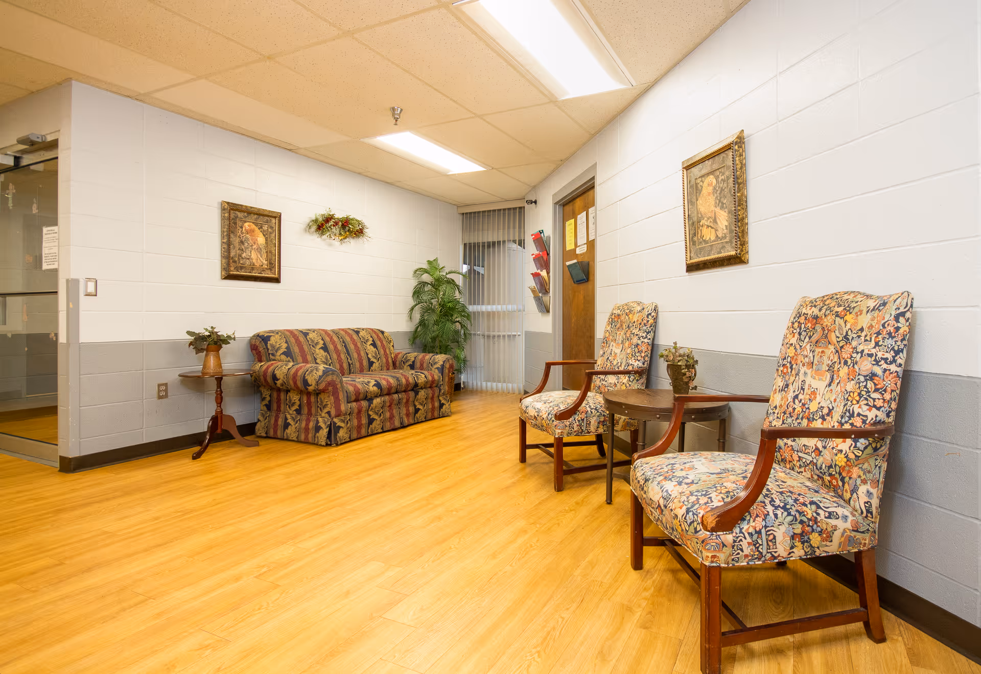 Seating area with patterned armchairs, a floral sofa, small tables, framed artwork, and wood flooring in a facility hallway.