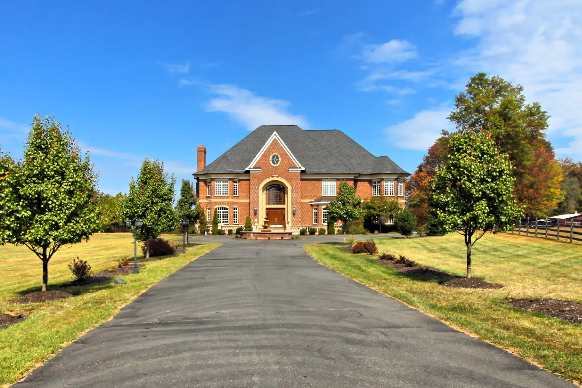 Front view of a large brick house at the end of a long paved driveway surrounded by lawns and trees.