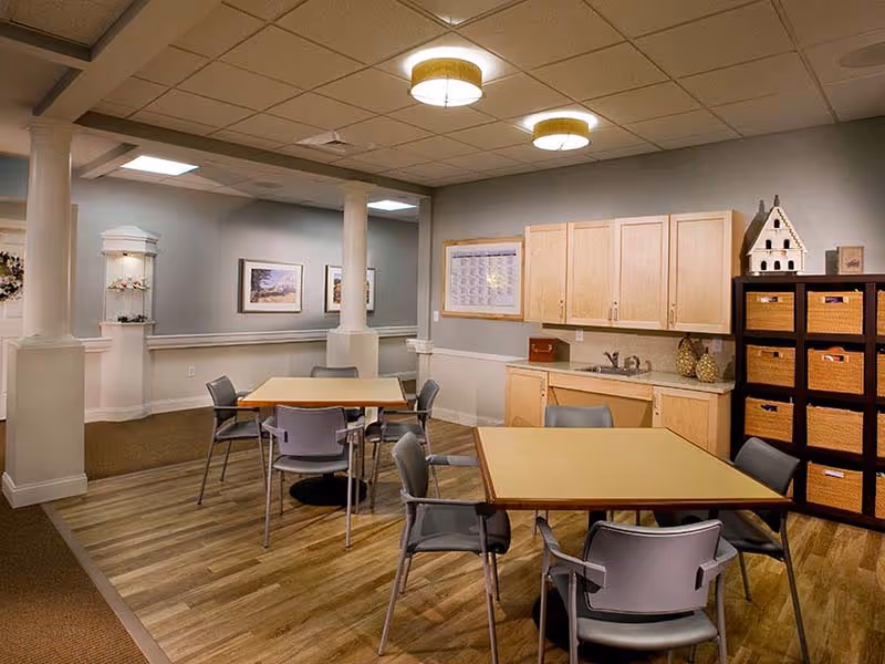 A well-lit common area with two square tables surrounded by gray chairs on a wood floor. The room features light-colored walls, two white columns, framed artwork, a small kitchenette with wooden cabinets and a sink, and a shelving unit with wicker baskets and decorative items.