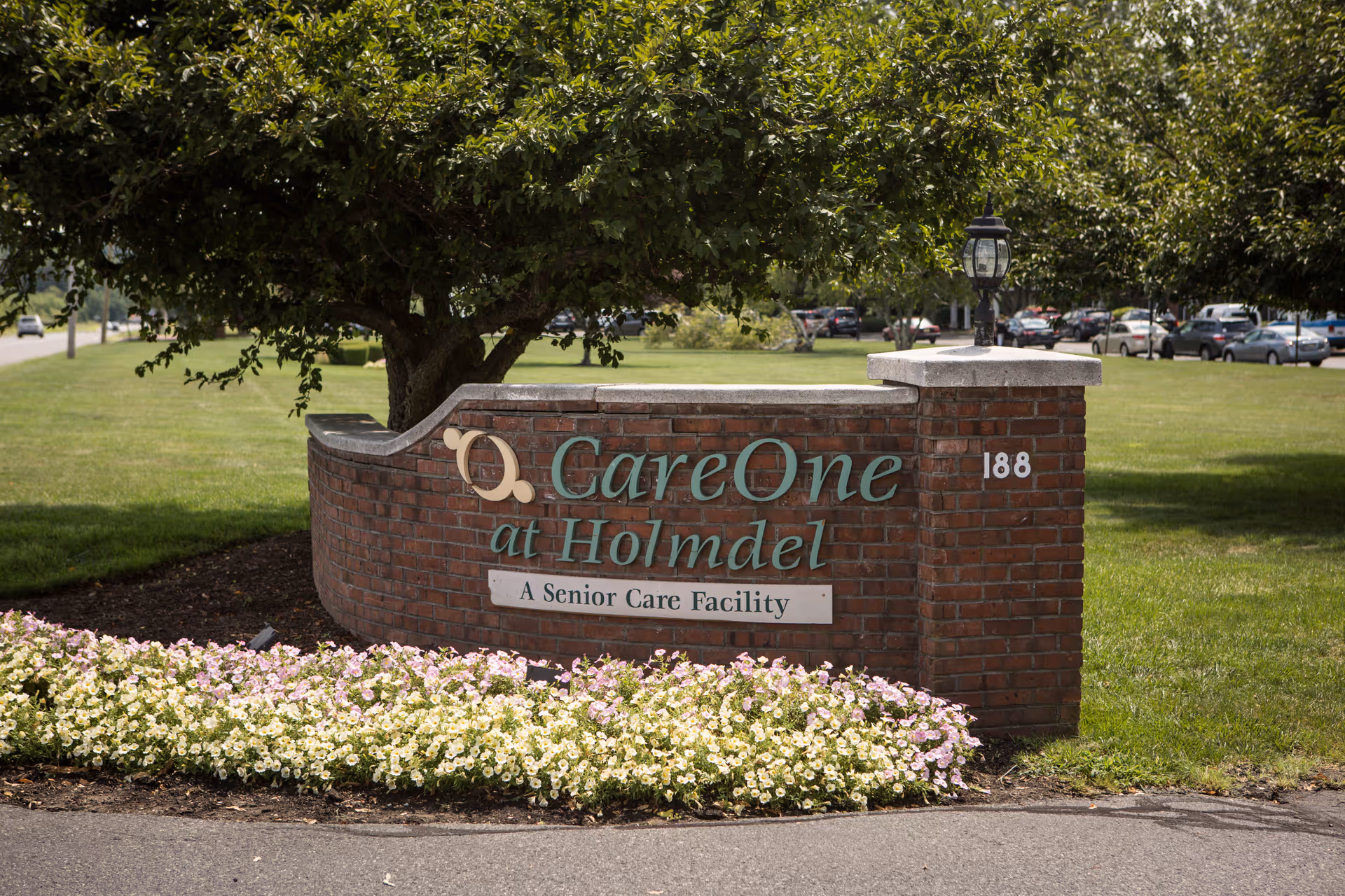 A brick sign for CareOne at Holmdel, a senior care facility, surrounded by green grass, a tree, and a flower bed with white and light purple flowers. The sign includes the address number 188 and a small lamp on top of the brick structure.