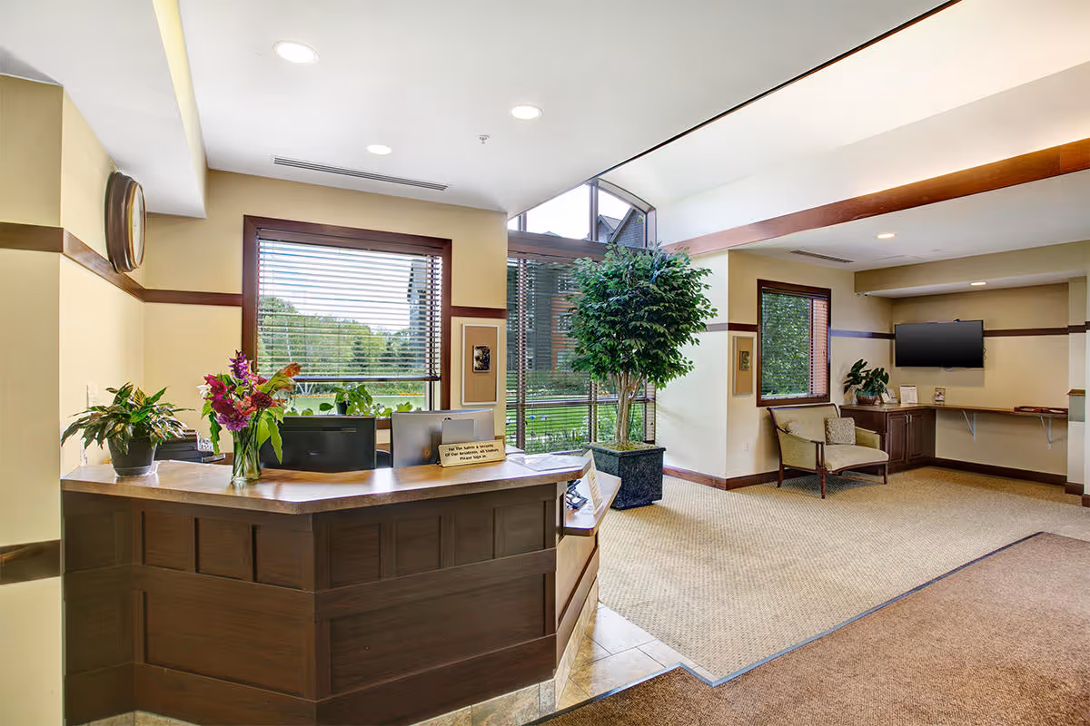 Reception area of Johanna Shores facility with a wooden front desk adorned with plants and flowers. Behind the desk are large windows with blinds, allowing natural light to brighten the space. To the right, there is a seating area with a chair, a mounted flat-screen TV, and a small counter with decorative items. The room has beige walls with wooden trim and a carpeted floor.