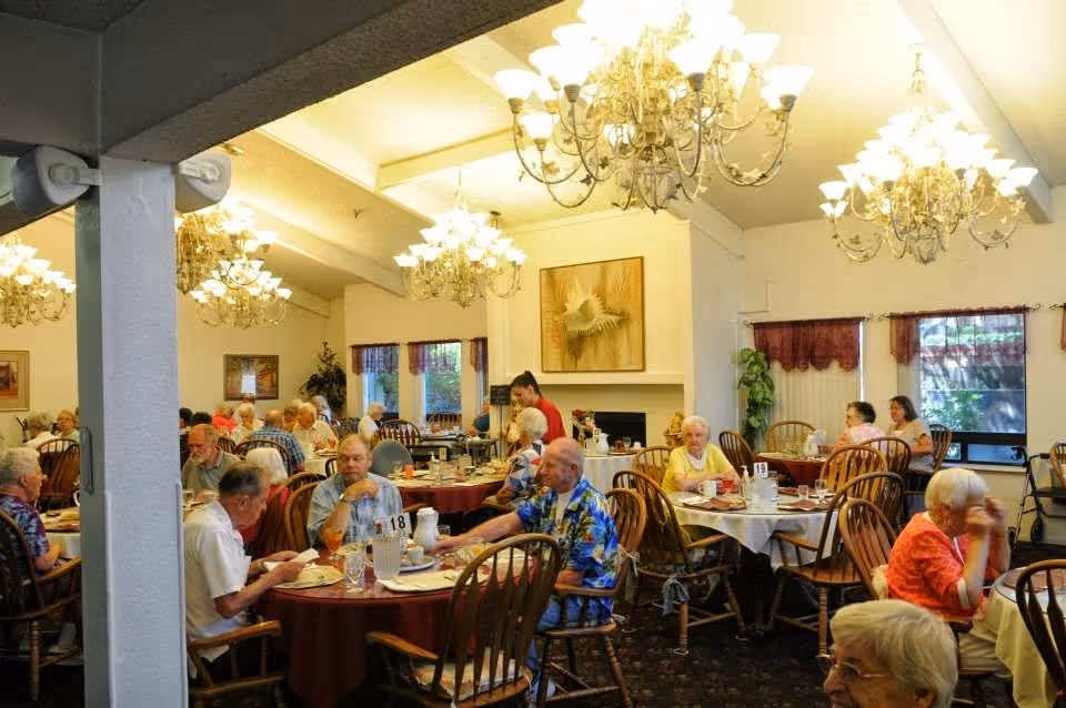 A busy dining room in a retirement facility with elderly residents seated at round tables covered with tablecloths, eating and socializing. The room is well-lit with multiple ornate chandeliers hanging from the ceiling. There are large windows with curtains, framed artwork on the walls, and a fireplace in the background. A staff member is attending to the residents.