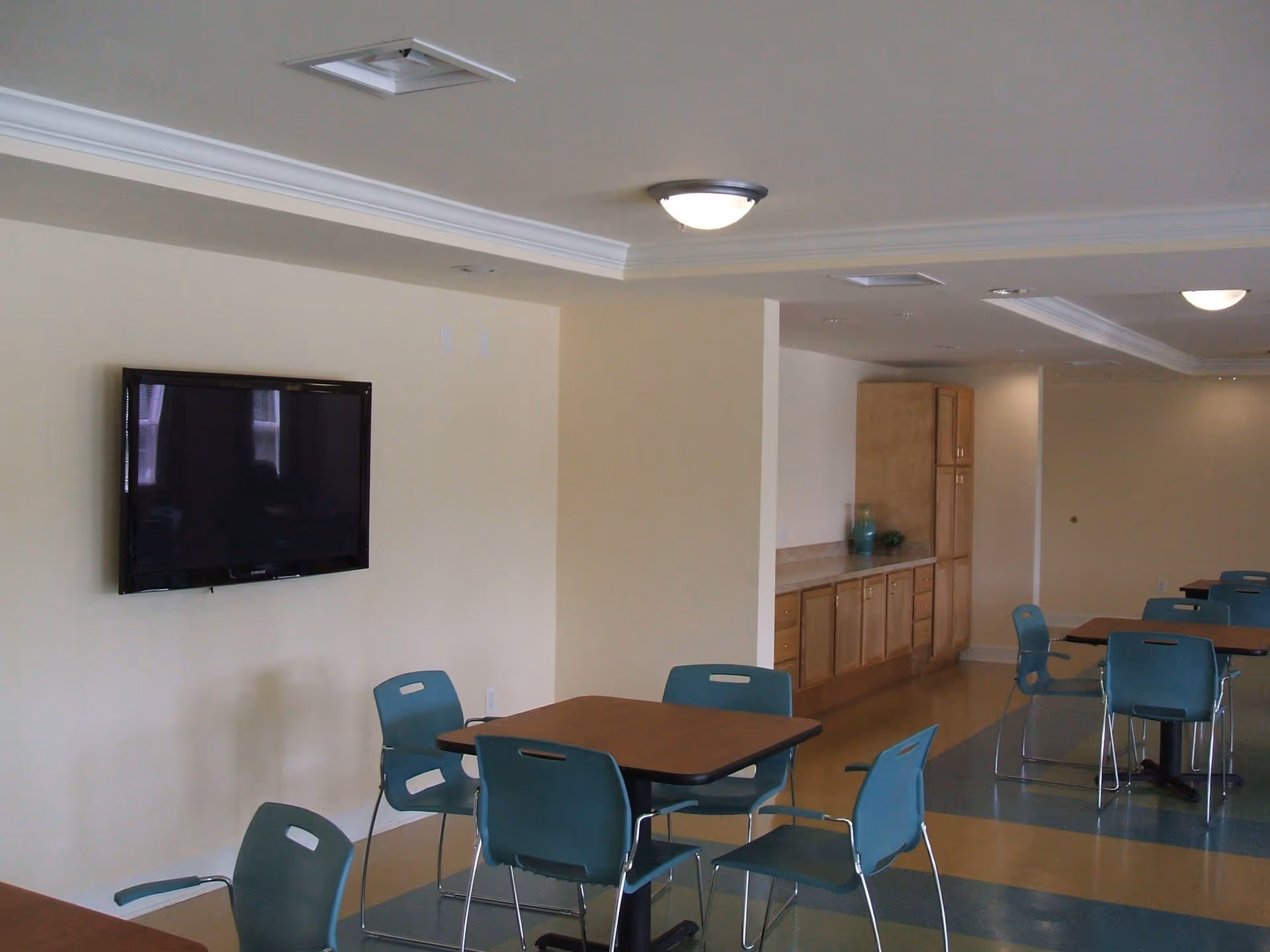 Interior view of a common area with several tables and blue chairs arranged around them. A flat-screen TV is mounted on a beige wall. There are wooden cabinets and a countertop along the back wall. The floor has a patterned design with blue and tan colors, and ceiling lights provide illumination.