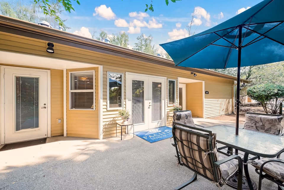 Patio with a glass-top table, umbrella and cushioned chairs in front of a yellow single-story building with French doors and windows.