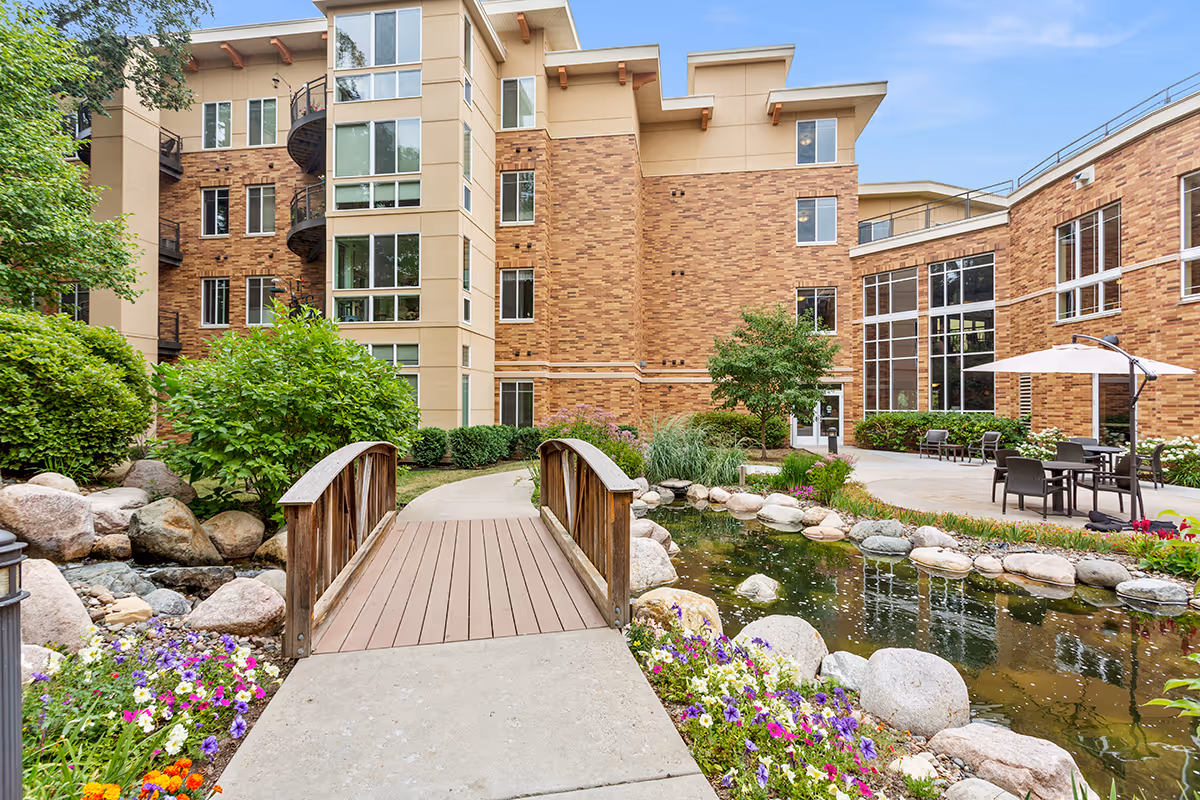 A landscaped courtyard featuring a small wooden footbridge over a pond, patio seating and umbrellas, colorful flowers, and a multi-story brick building in the background.