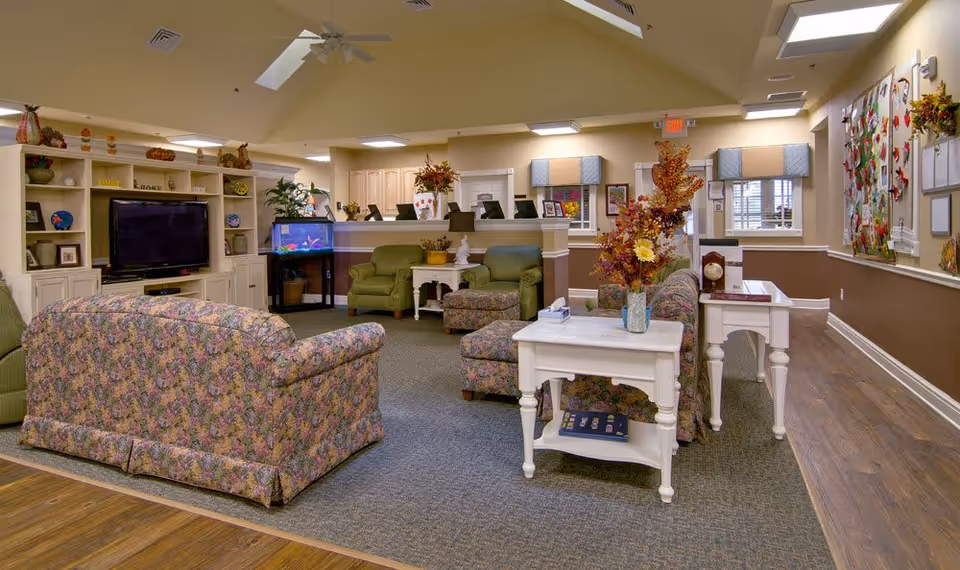 A cozy living room area in a senior living facility with floral patterned sofas and armchairs, a white coffee table with a vase of flowers, a TV on a white entertainment center, an aquarium, and a reception desk in the background. The walls are decorated with artwork and bulletin boards, and the room has a warm, inviting atmosphere.