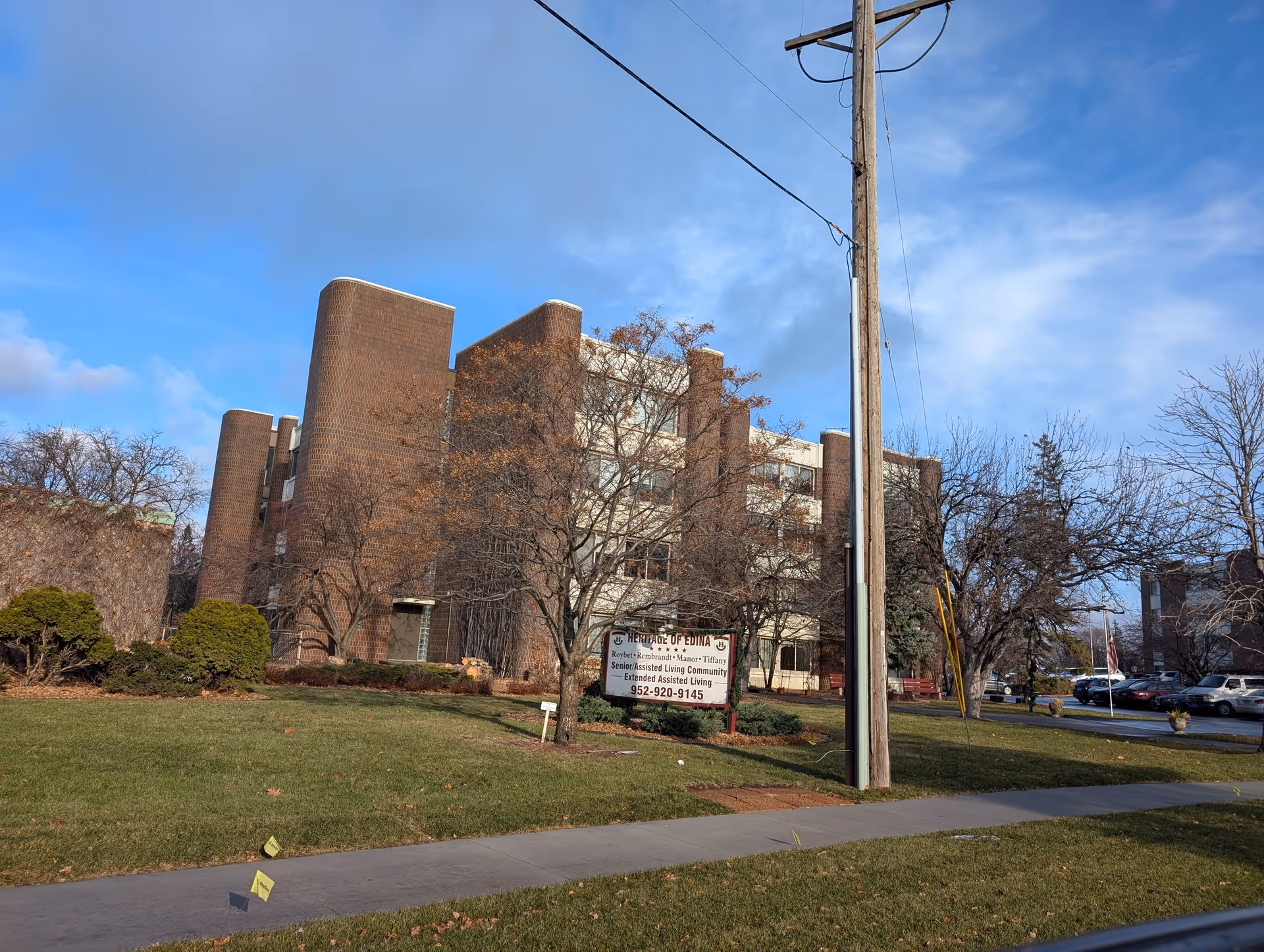 Brick multi-story senior living building with a lawn, trees, a front sign and a utility pole under a blue sky.