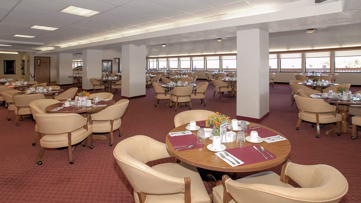 Spacious dining room with multiple round tables set with cups, glasses, silverware, and napkins. Each table has beige cushioned chairs and a small floral centerpiece. The room has large windows along one wall, carpeted floors, and white ceiling panels with fluorescent lighting.