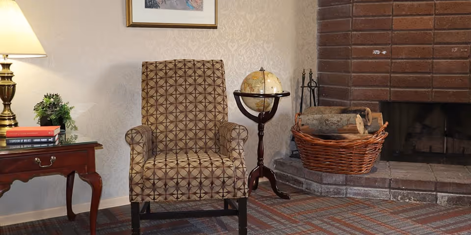 A cozy living room corner featuring a patterned armchair, a wooden side table with a lamp and books, a globe on a stand, and a brick fireplace with a basket of firewood.