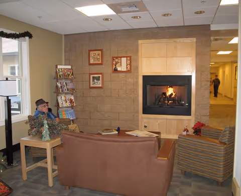 Cozy common-room lounge with chairs and a couch facing a lit fireplace, a magazine rack, and a person seated near a window.