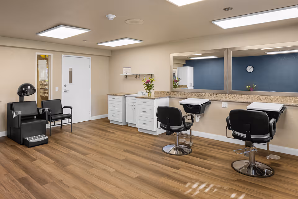 Interior view of a senior living facility's hair salon area with two black salon chairs in front of mirrors, a hair washing station, a black hair dryer chair, a small black waiting chair, white cabinets with a granite countertop, and a wooden floor.