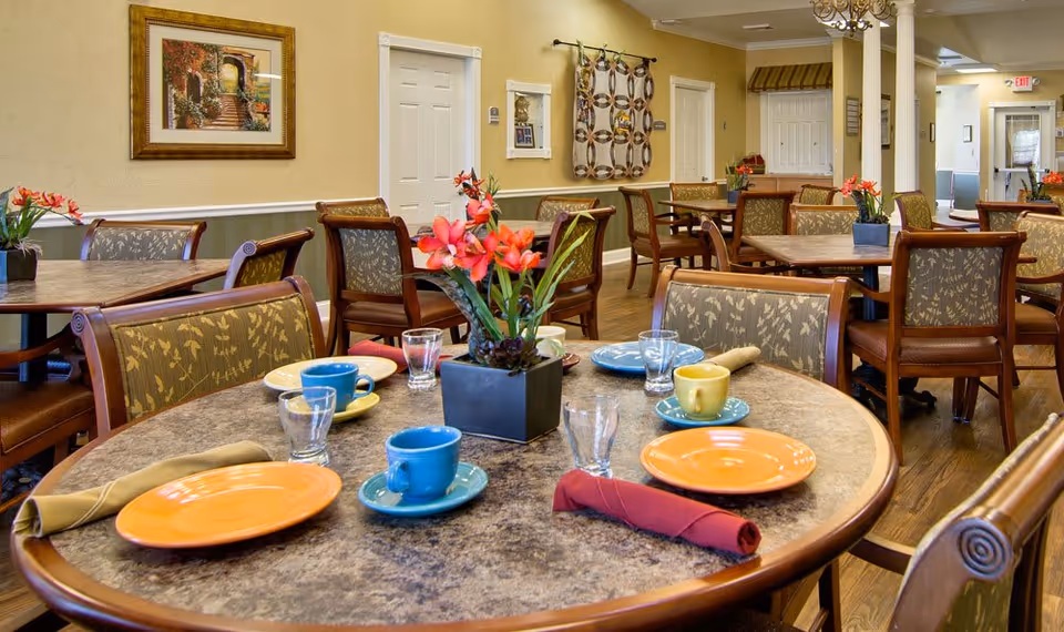 A dining room in a senior living facility with round tables set with colorful plates, cups, glasses, and napkins. Each table has a small flower arrangement in the center. The room has wooden chairs with patterned upholstery, light yellow walls, framed artwork, and a wooden floor.