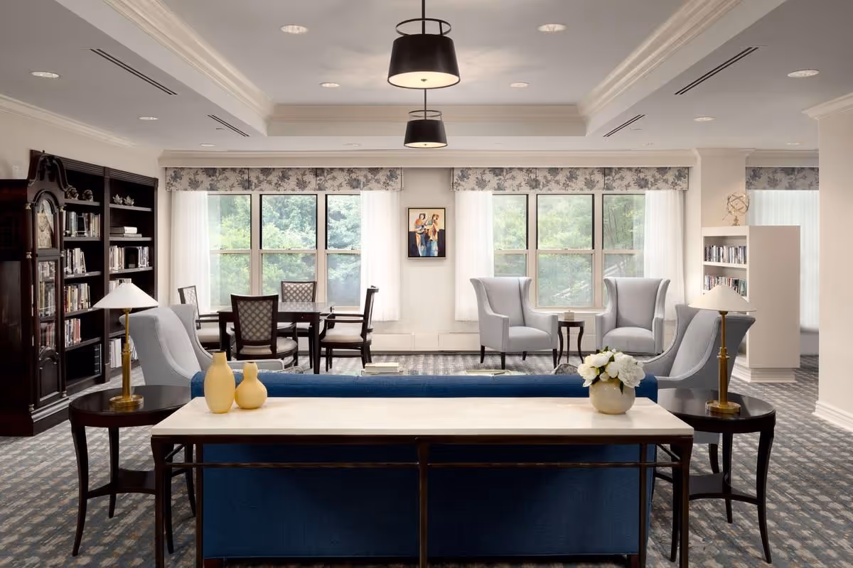 Bright, elegant communal living room with sofas, armchairs, bookshelves and a dining table by large windows.