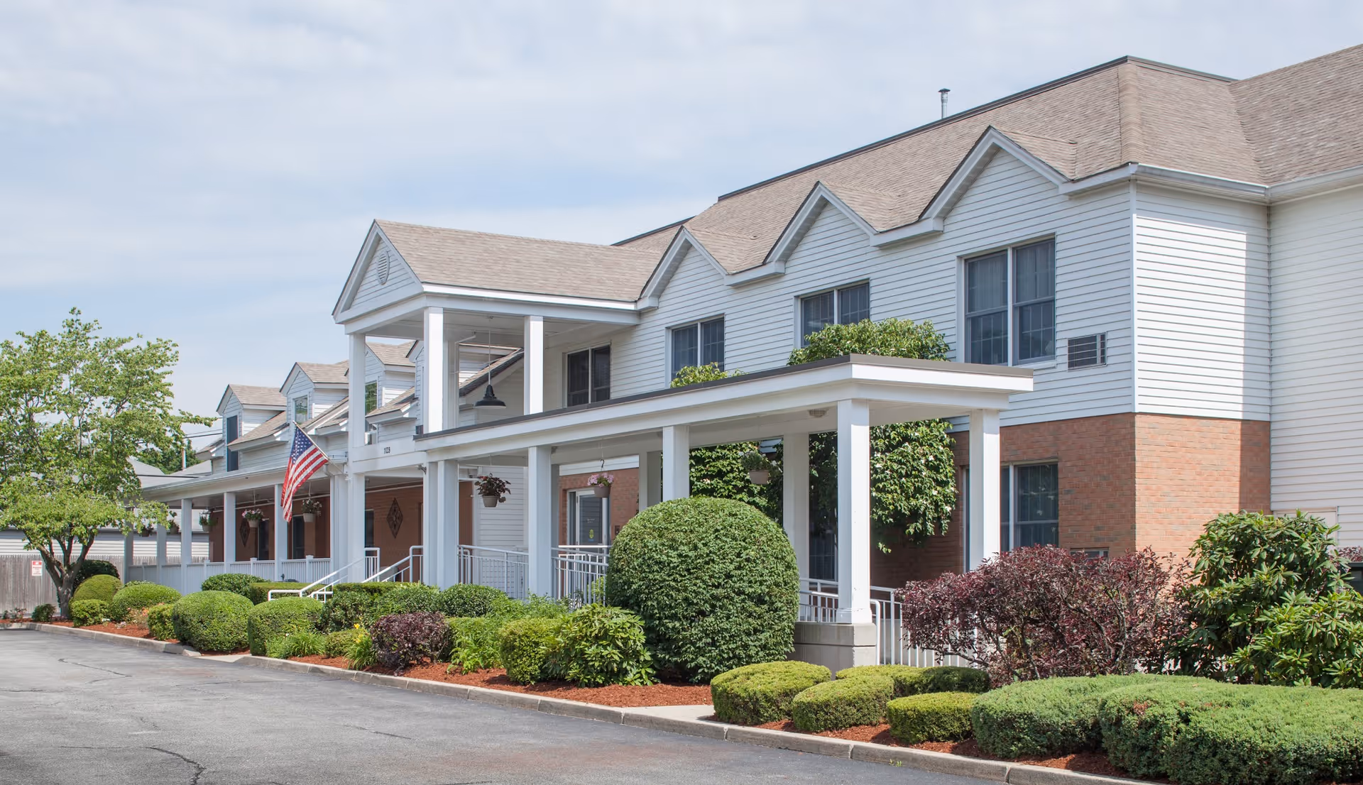 Exterior view of a two-story senior living facility building with white siding and brick accents. The building features a covered porch with white columns, neatly trimmed bushes, and an American flag near the entrance. The sky is partly cloudy.