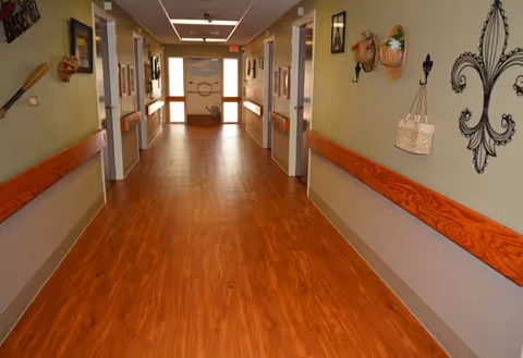 A long, clean hallway in a nursing and rehabilitation center with wood-patterned flooring and handrails on both sides. The walls are decorated with various wall hangings, including baskets and ornamental designs. Doors line both sides of the hallway, and natural light comes from windows at the far end.
