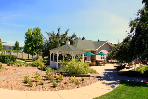 Outdoor garden area at Juniper Village at Aurora featuring a white gazebo surrounded by plants and flowers, paved walkways, patio tables with green umbrellas, and a building in the background under a clear blue sky.