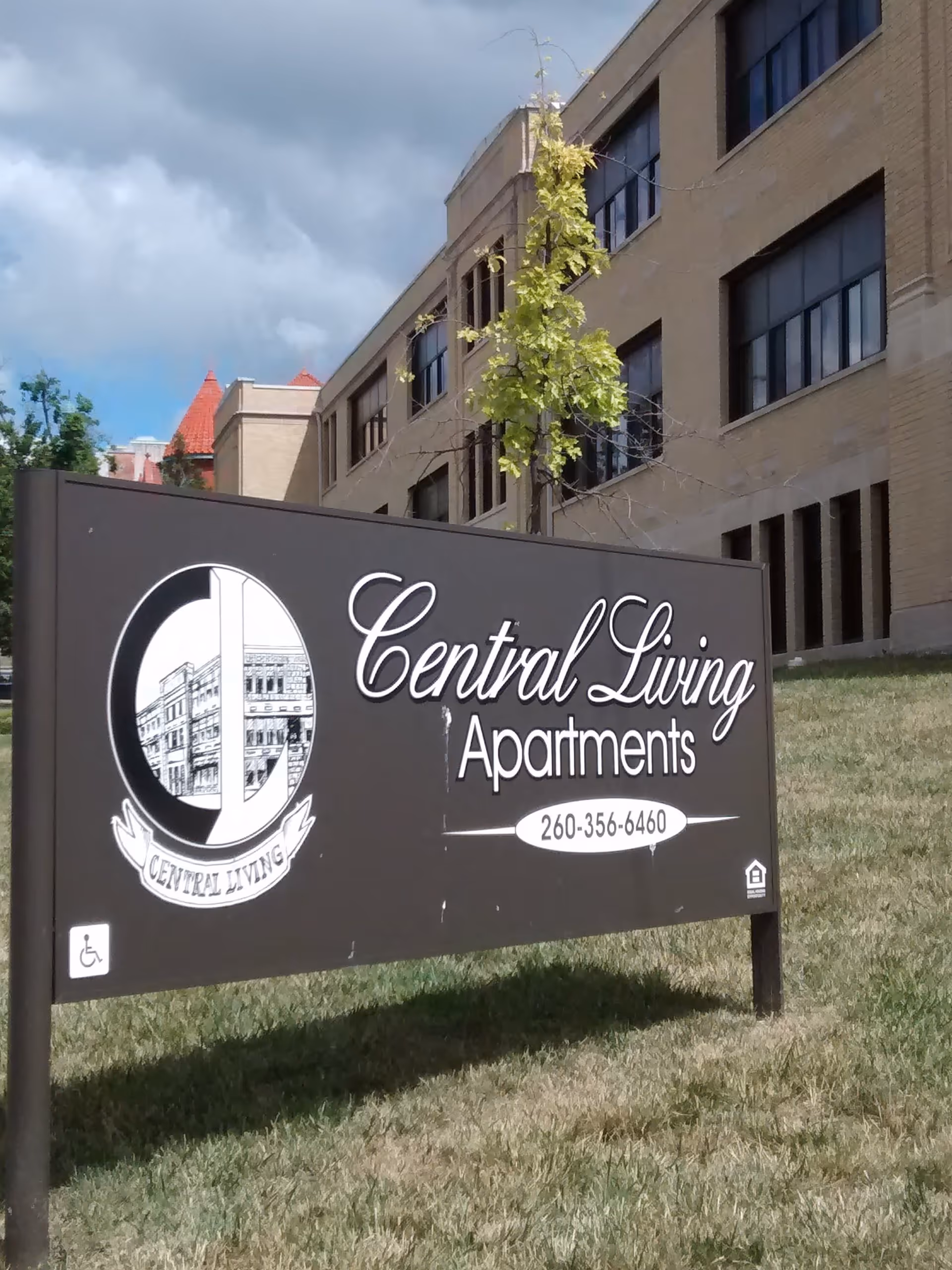 A black sign on a grassy area in front of a beige brick building. The sign reads 'Central Living Apartments' with a phone number 260-356-6460 and includes a logo with a building illustration. There is a small tree behind the sign and a partly cloudy sky above.