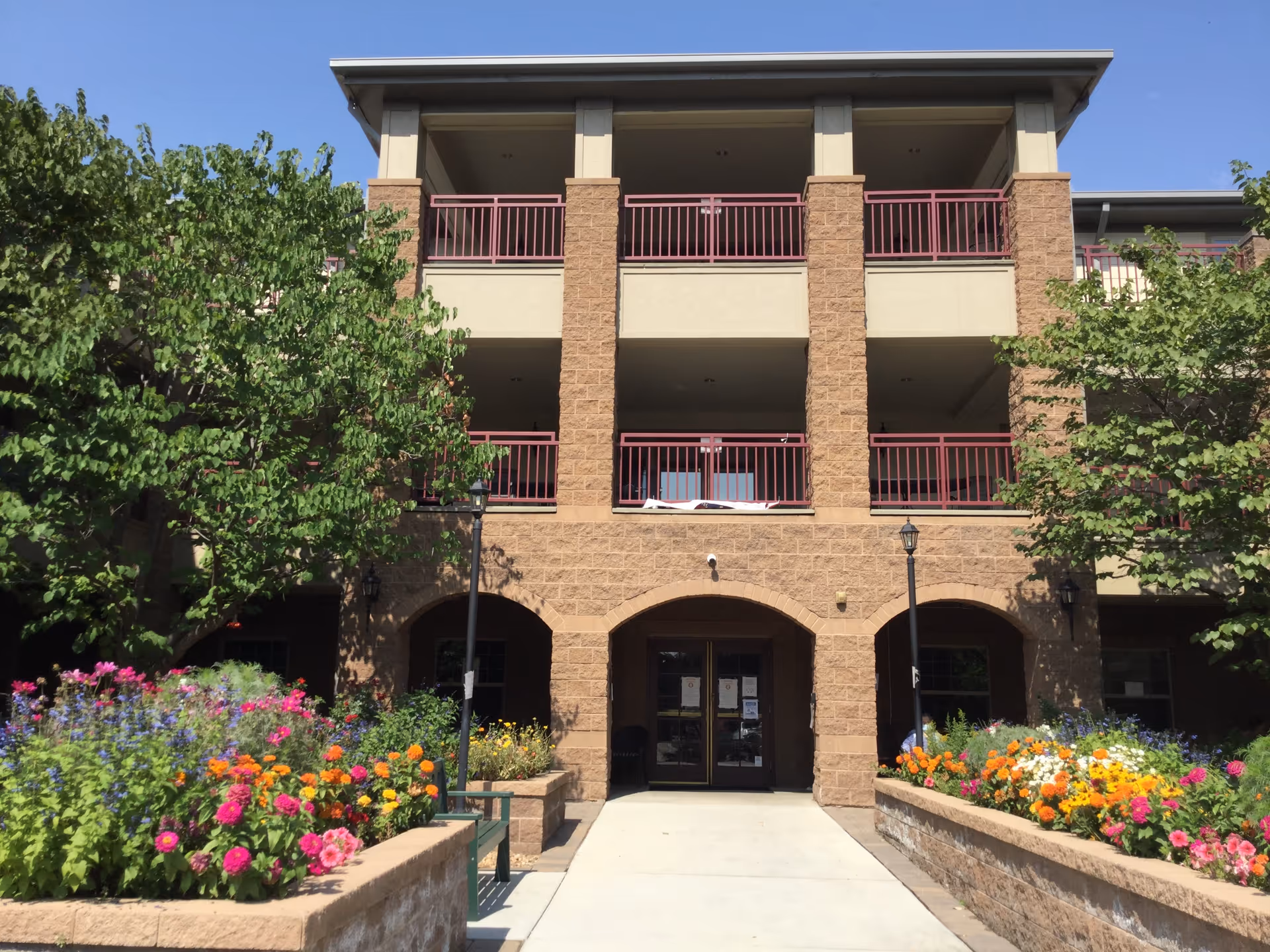 Front exterior view of a multi-story assisted living center building with stone and beige walls, red railings on balconies, and a walkway leading to the entrance. There are colorful flower beds and green trees on either side of the walkway under a clear blue sky.
