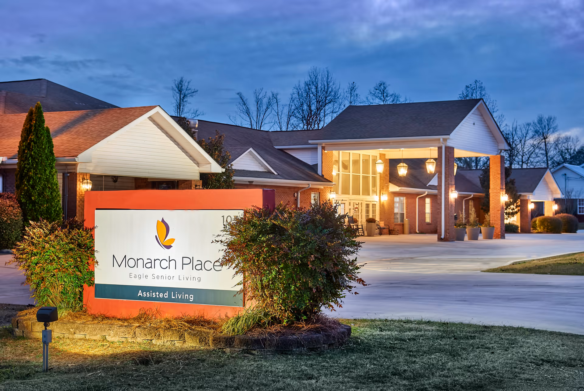 Exterior view of Monarch Place assisted living facility during dusk, showing a well-lit entrance with a covered driveway and a large sign in front surrounded by bushes and landscaping.