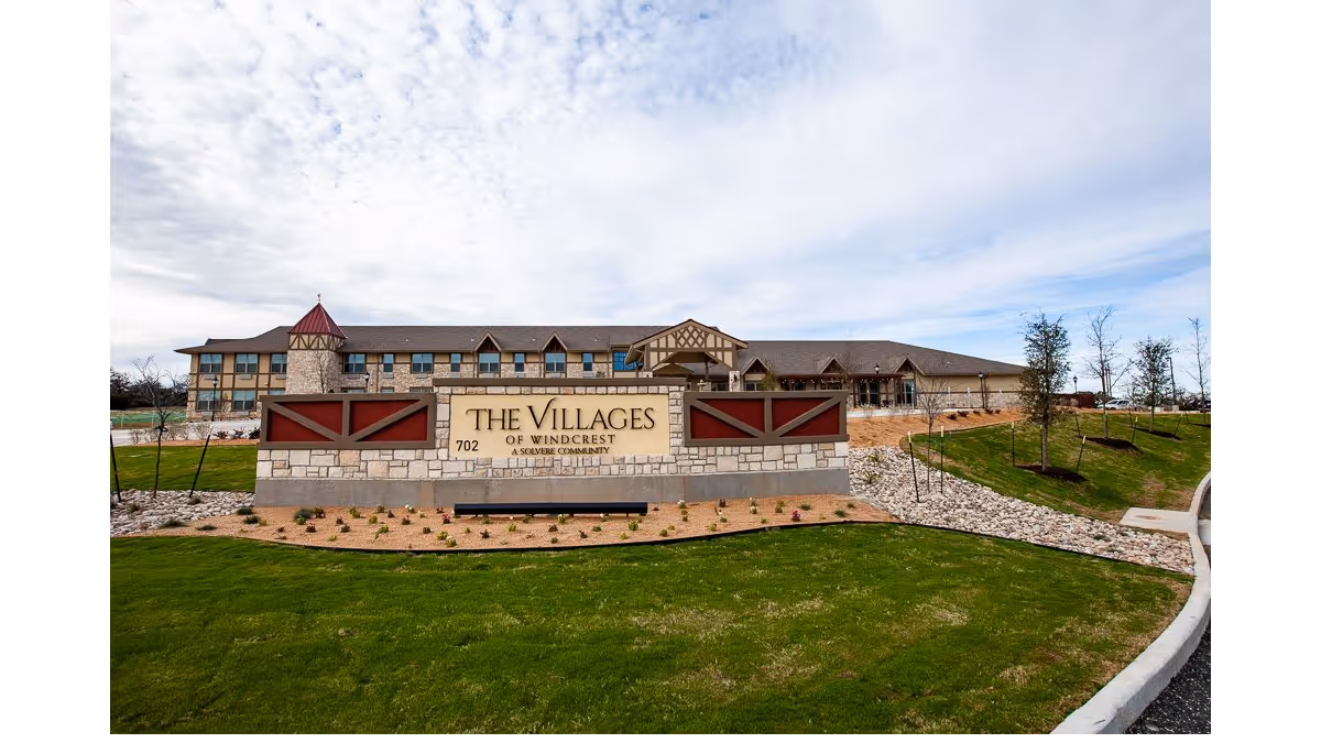 Exterior view of The Villages of Windcrest senior living facility with a large stone sign in the foreground displaying the facility name and address. The building has a multi-story design with a turret and pitched roofs, surrounded by landscaped grass, small plants, and young trees under a partly cloudy sky.