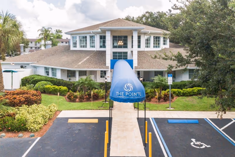 Front exterior view of The Pointe senior living community building with a blue canopy entrance, surrounded by landscaped greenery and parking spaces including a handicapped spot.
