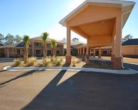 Exterior view of a senior living facility with a covered entrance supported by brick columns, a paved driveway, landscaped area with small palm trees and shrubs, and a two-story building in the background under a clear sky.