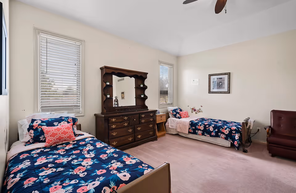 Bright shared bedroom with two twin beds, floral bedding, a wooden dresser with mirror, and a armchair.