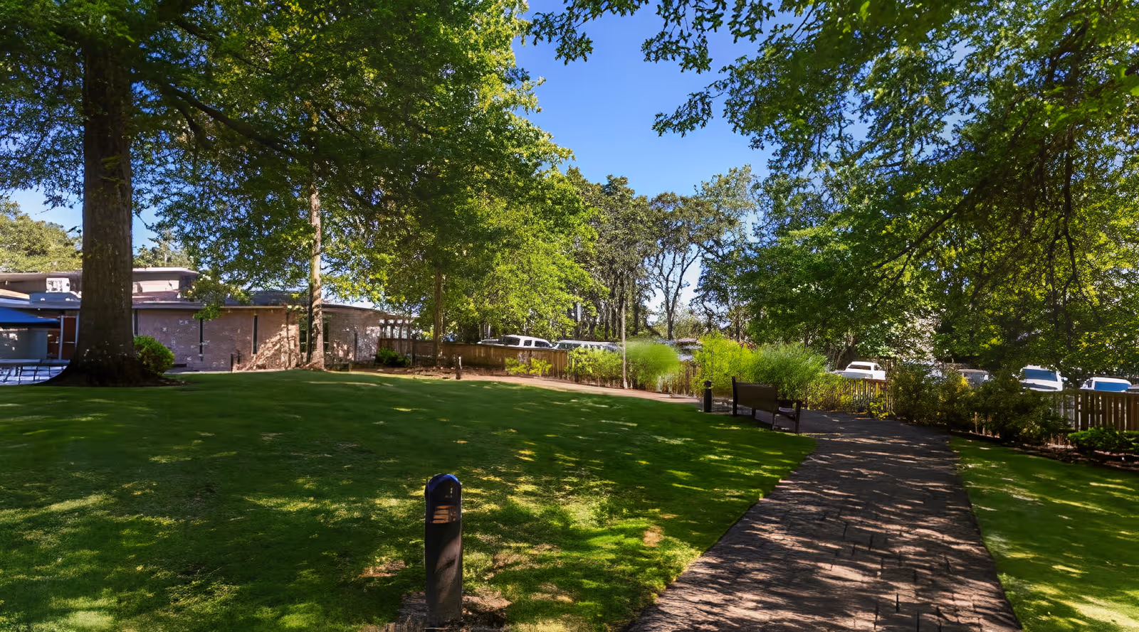 A peaceful outdoor garden area at Hillside Heights Rehabilitation Center with a paved walking path, green grass, large trees providing shade, benches along the path, and a building partially visible in the background under a clear blue sky.