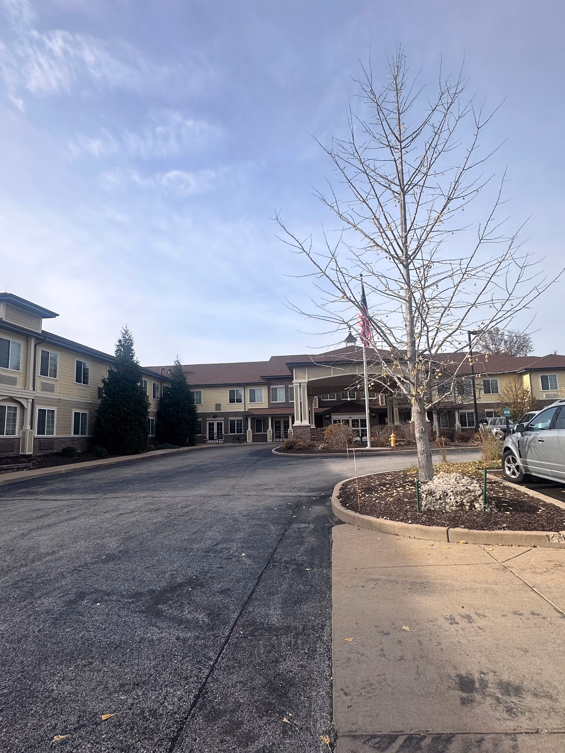 Two-story assisted living building with a circular driveway, a leafless tree and flagpole in front under a cloudy sky.