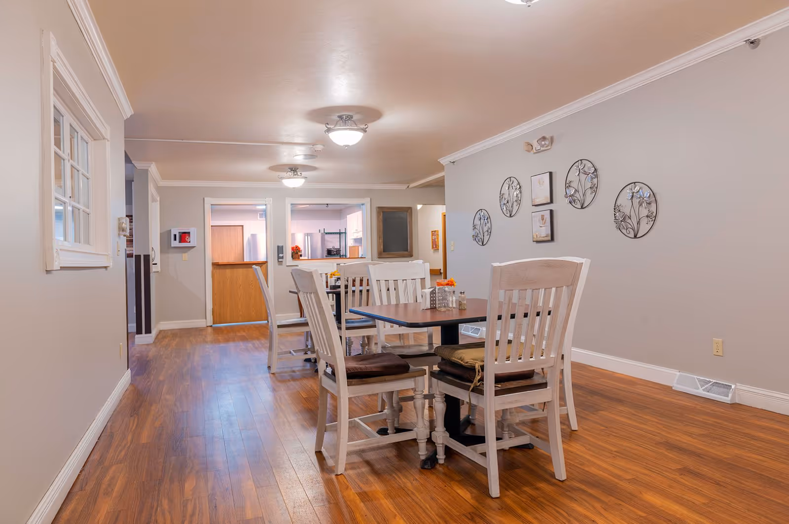 A dining area in a senior living facility with wooden flooring, a table with four white wooden chairs, and wall decorations including metal floral art and framed pictures. The room is well-lit with ceiling lights and has a view into an adjacent kitchen area.
