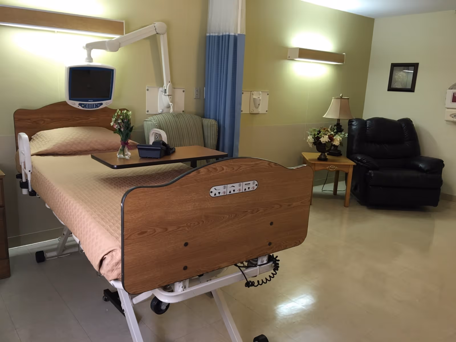 A hospital-style room with a medical bed covered in a beige quilt, a mounted monitor above the bed, a small table with a telephone and a vase of flowers, a striped armchair partially visible behind a blue and white privacy curtain, a wooden side table with a lamp and a flower arrangement, and a black recliner chair against a light green wall.