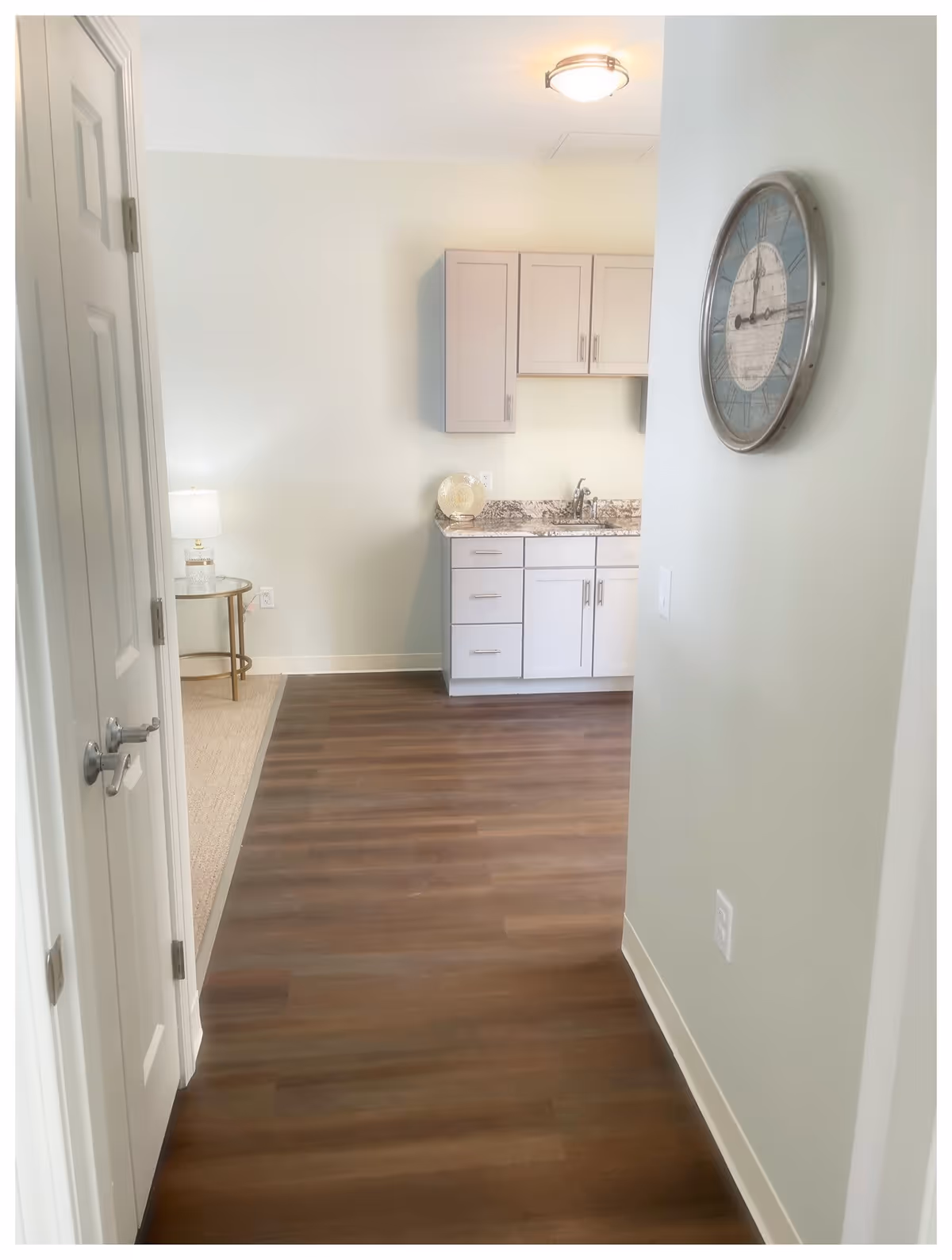 View down a hallway with wood flooring leading to a small kitchenette area with light-colored cabinets and a granite countertop. A round wall clock is mounted on the right wall, and a small side table with a lamp is visible in an adjacent carpeted room on the left.