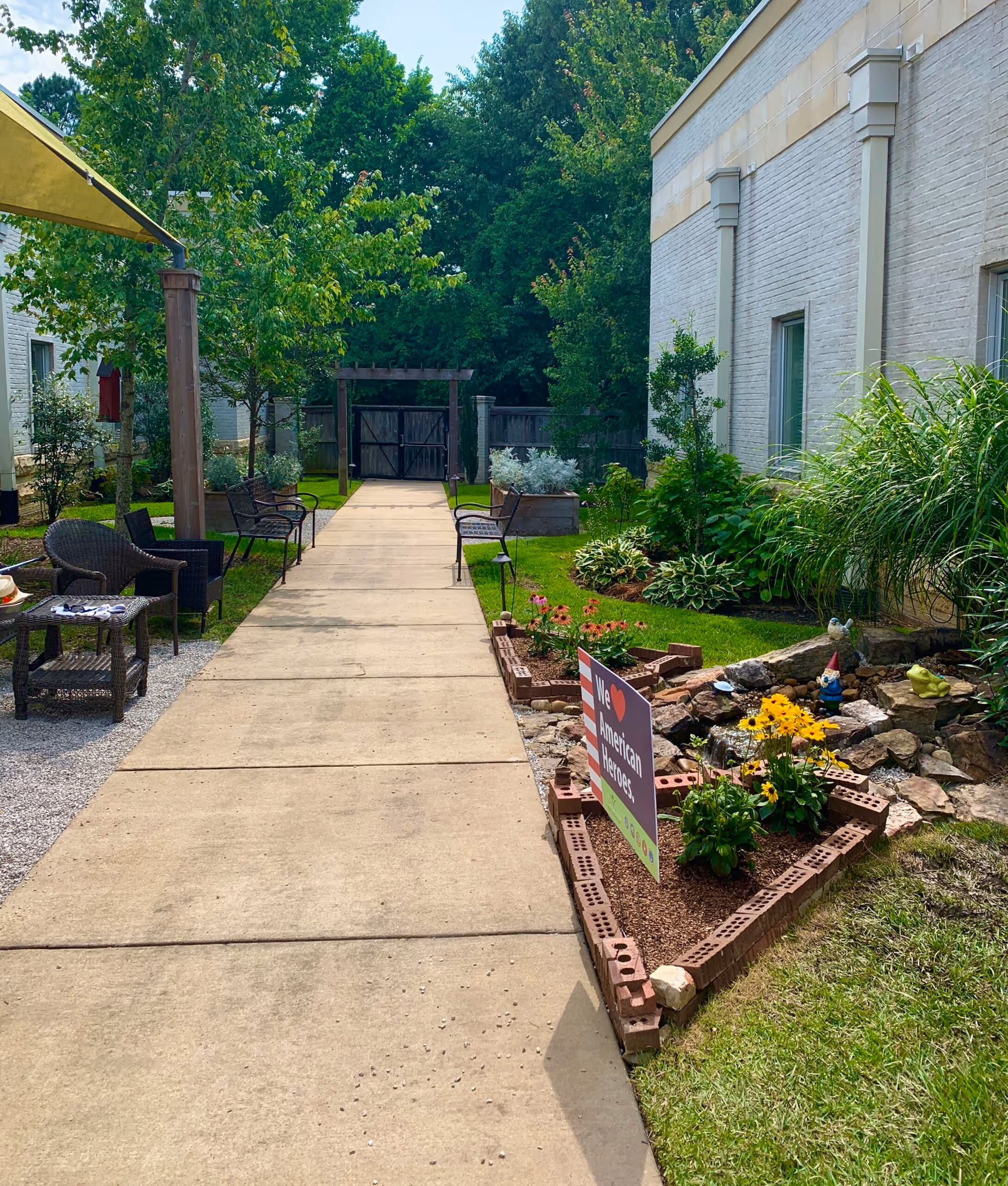A paved walkway flanked by garden beds with flowers and greenery on the right and outdoor seating with chairs and a table on the left. The area is surrounded by trees and a white brick building. A sign in the garden bed reads 'We ♥ American Heroes.'