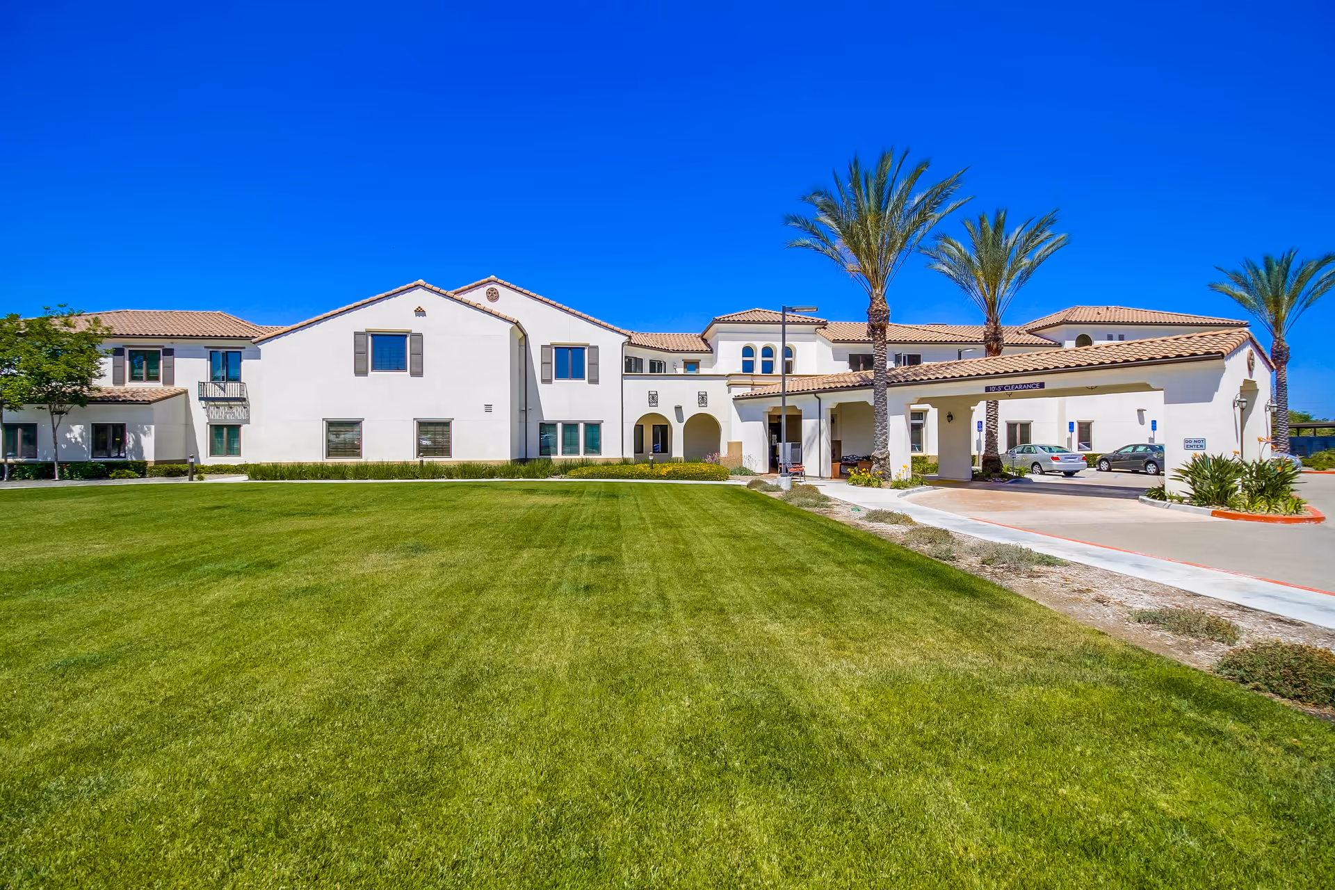 Exterior view of a senior living facility building with white walls and a tiled roof, surrounded by a well-maintained green lawn and palm trees under a clear blue sky. There is a driveway with parked cars near the entrance.