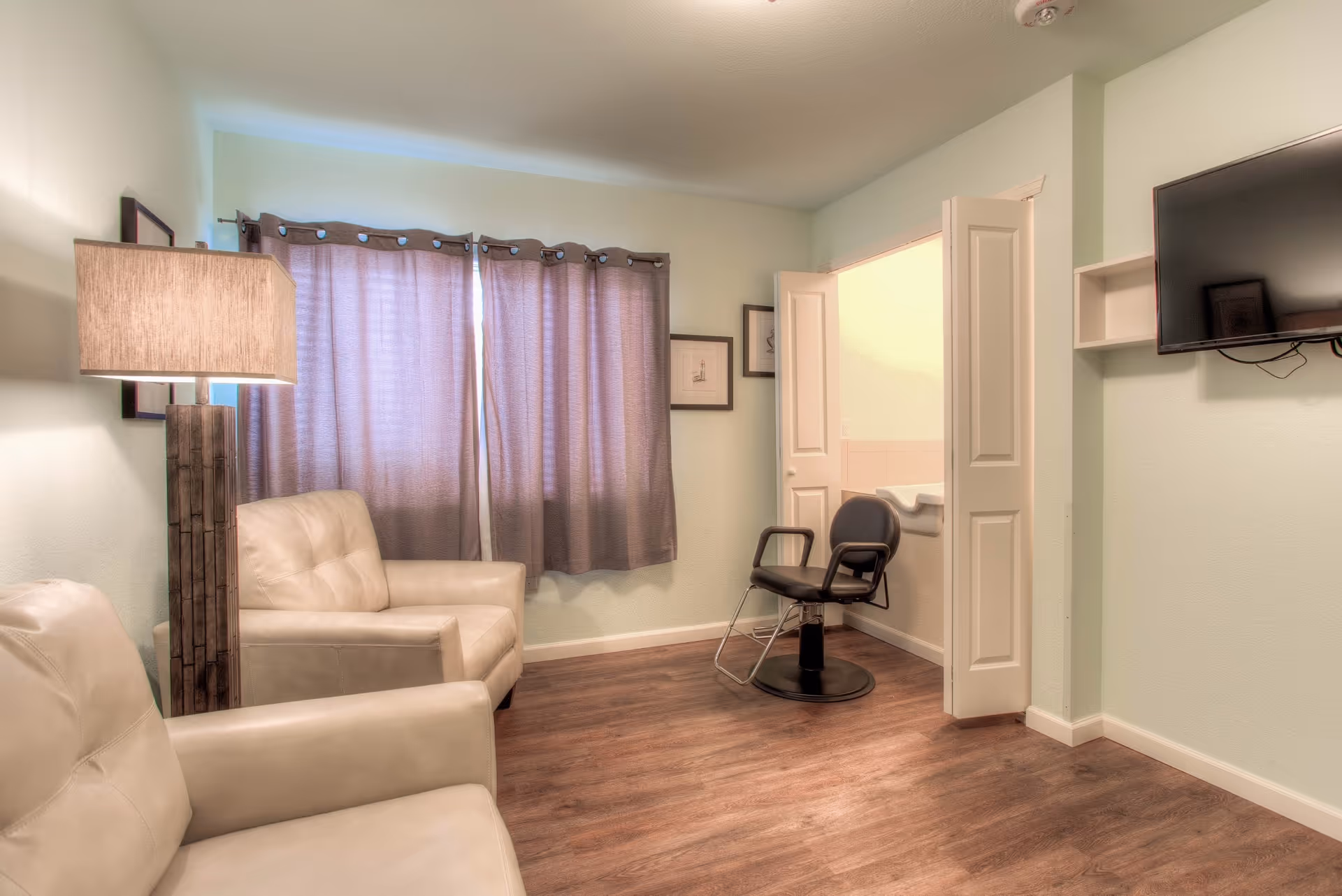 A cozy room with two beige leather armchairs, a tall wooden floor lamp, and a window with closed purple curtains. There is a black salon chair in an adjacent small room visible through open double doors. A flat-screen TV is mounted on the wall to the right.
