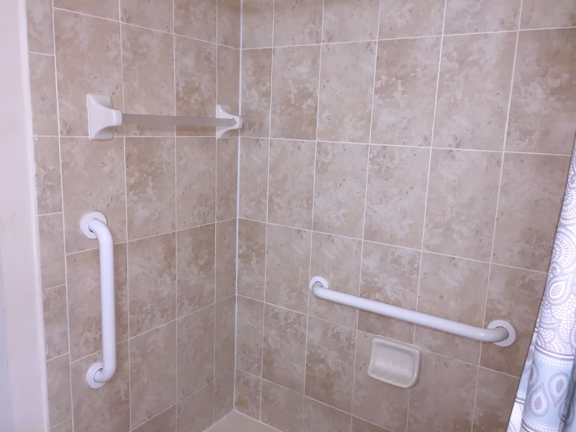 Corner of a bathroom shower area with beige tiled walls, featuring two white grab bars, a towel bar, a soap holder, and a partially visible patterned shower curtain on the right side.