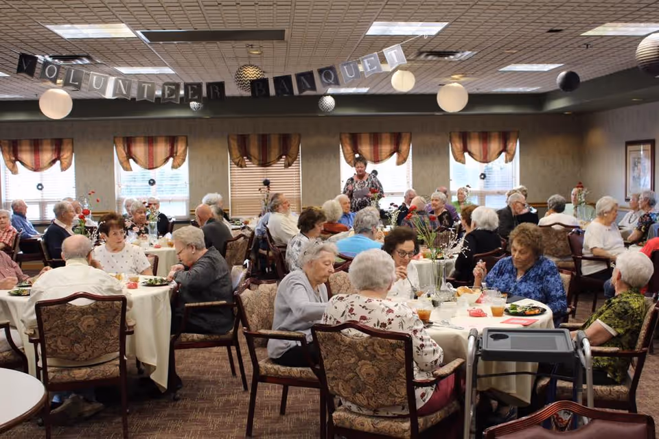 A large group of elderly people seated at round tables in a decorated dining room, enjoying a meal together. The room has multiple windows with striped valances, and a banner hanging from the ceiling reads 'VOLUNTEER BANQUET'. The atmosphere appears social and communal.