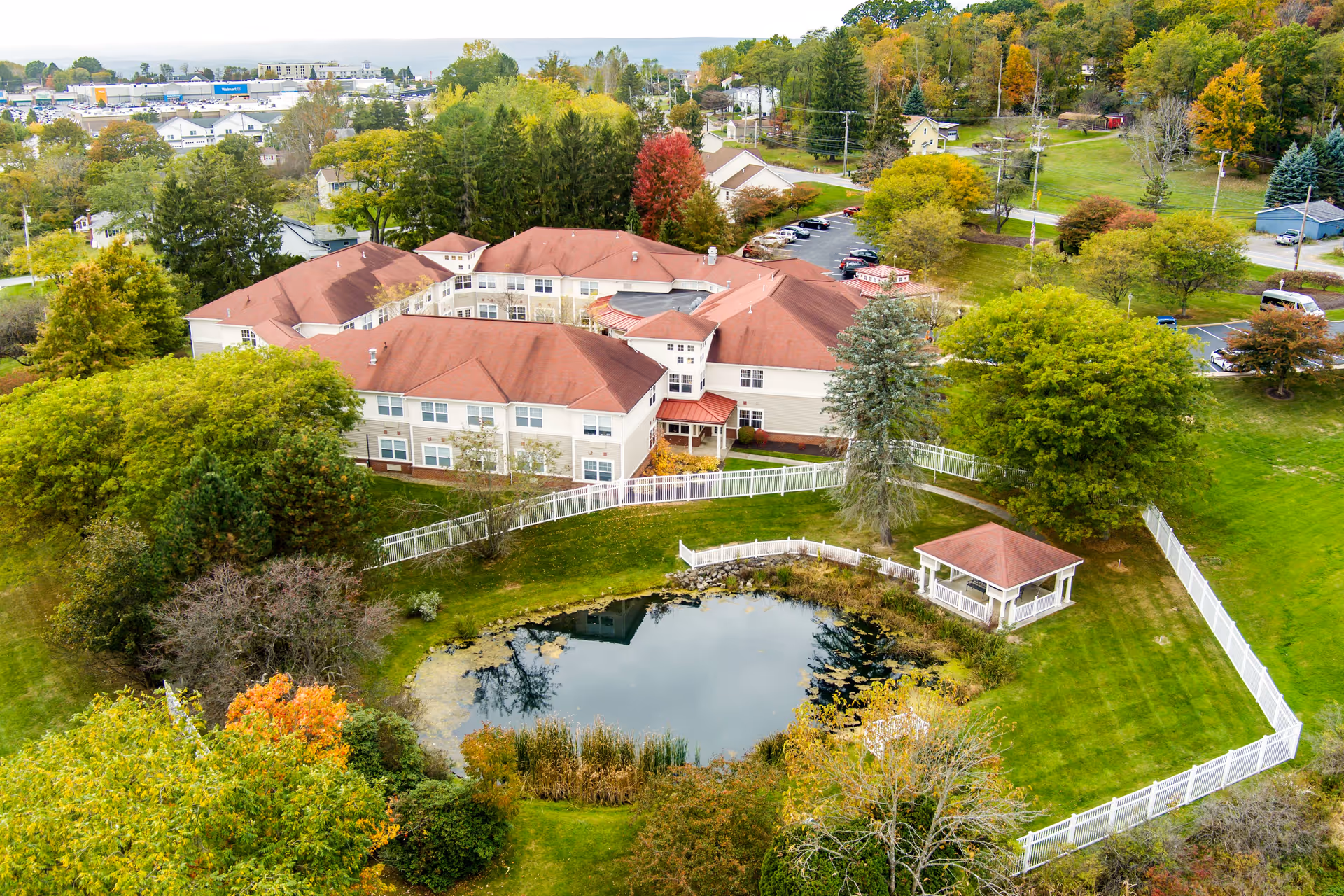 Aerial view of a red-roofed senior living complex with a white fence, small pond and gazebo surrounded by trees and lawns.