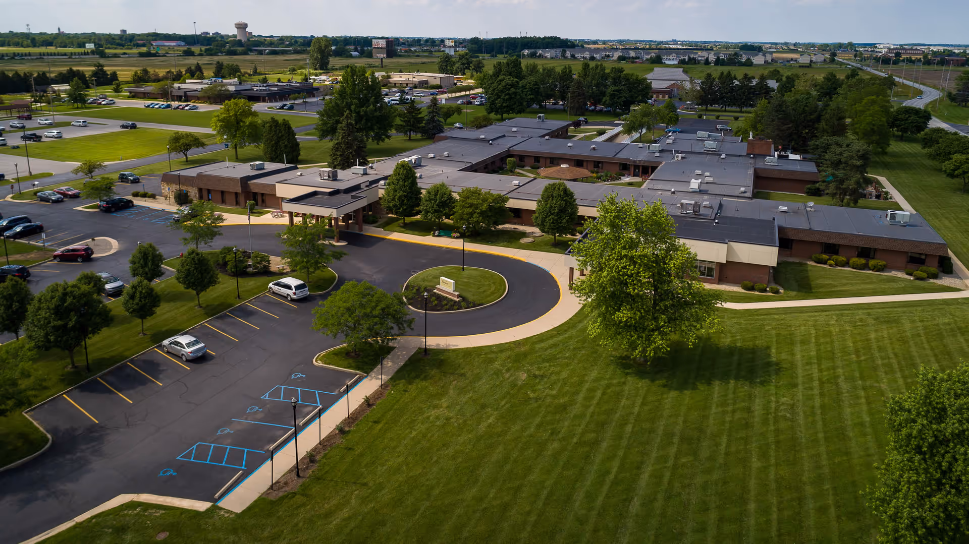 Aerial view of Wood Haven Healthcare facility showing a large single-story building surrounded by well-maintained green lawns, trees, and parking lots with several cars parked. The building has a circular driveway entrance and is located in a suburban area with other buildings and open fields in the background.
