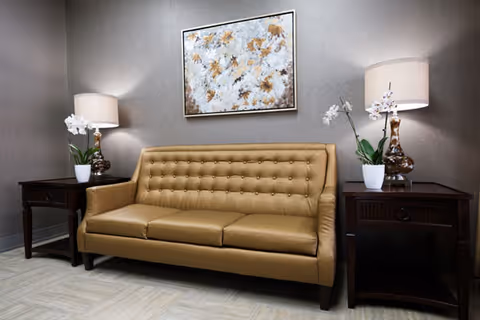 A beige leather tufted sofa is centered against a gray wall with a framed abstract floral painting above it. On each side of the sofa is a dark wooden side table, each holding a white orchid plant and a table lamp with a beige shade. The floor has light-colored tiles.