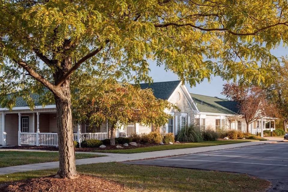 Single-story senior living building with white porches, landscaped shrubs, a large tree in the foreground, and a parking area.