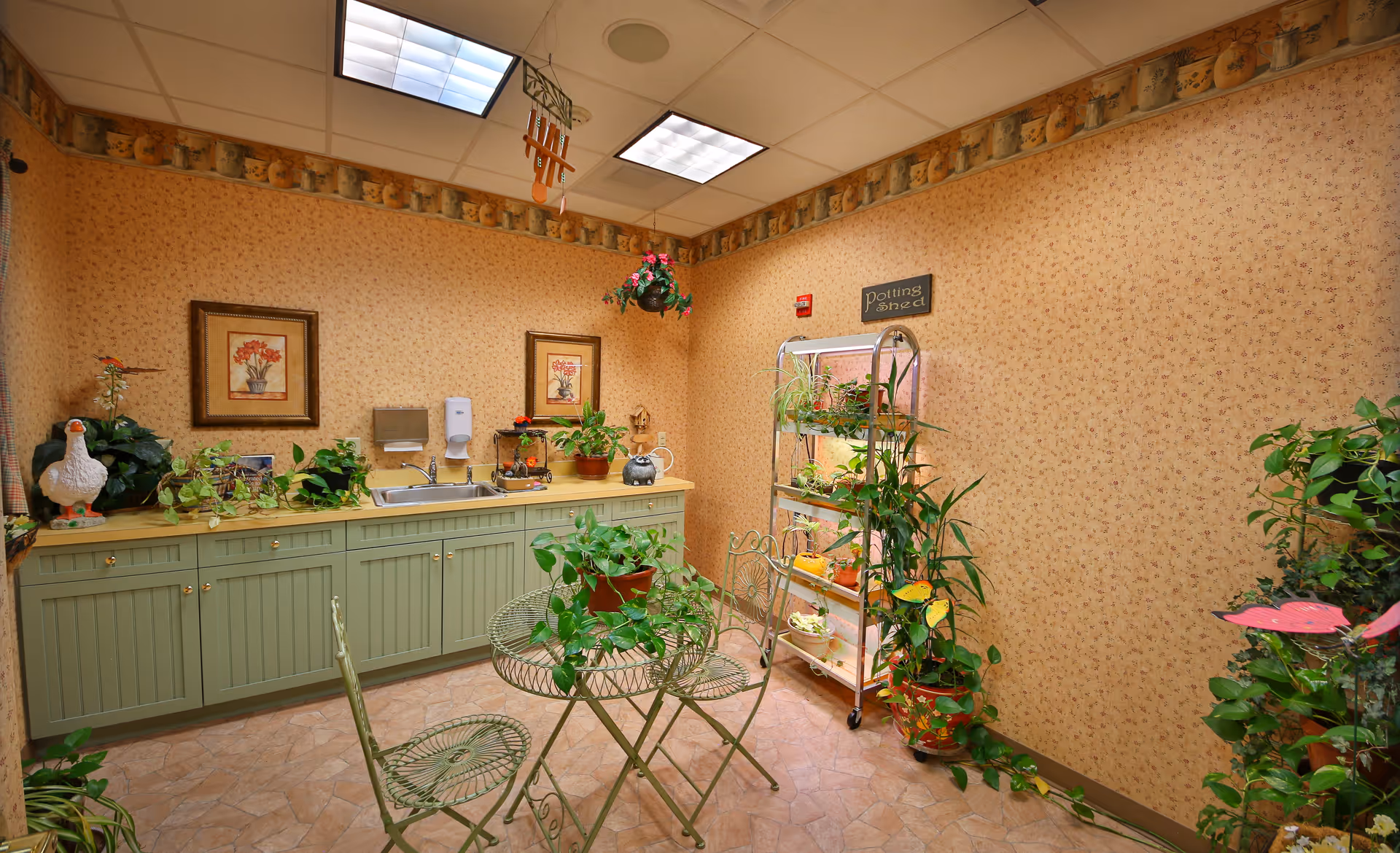 A cozy indoor potting area with green cabinets, a sink, and various potted plants on the countertop and shelves. There is a small round metal table with two matching chairs in the center, surrounded by more plants. The walls are decorated with floral wallpaper and framed pictures, and a sign above the shelves reads 'Potting Area'.