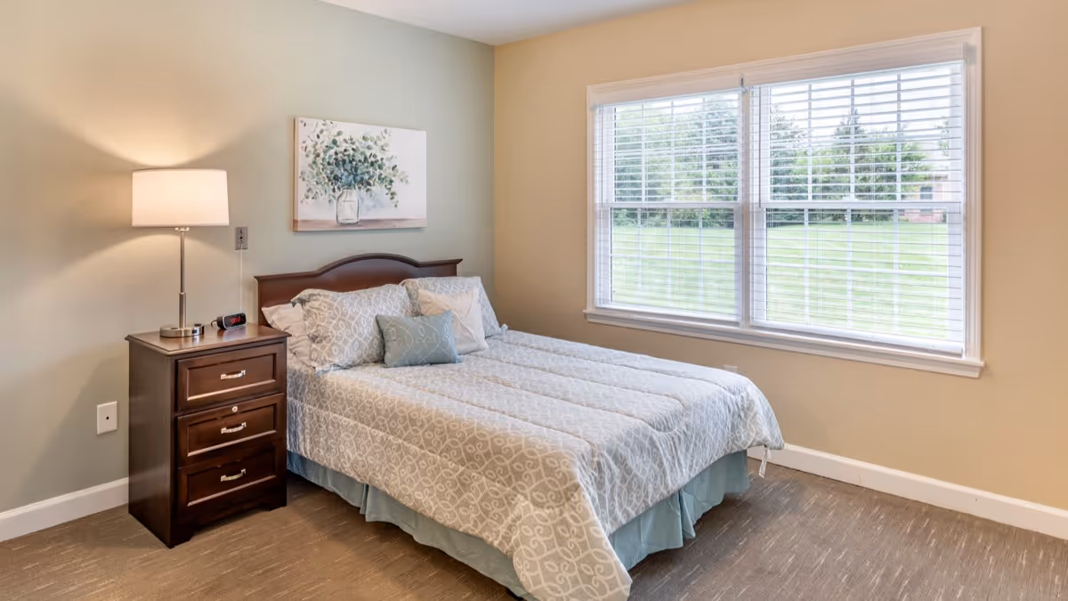 A bedroom with a neatly made bed featuring patterned bedding and multiple pillows. Next to the bed is a dark wooden nightstand with three drawers, a lamp, and a digital clock. A large window with white blinds lets in natural light, and a framed artwork of a vase with flowers hangs on the wall above the bed.