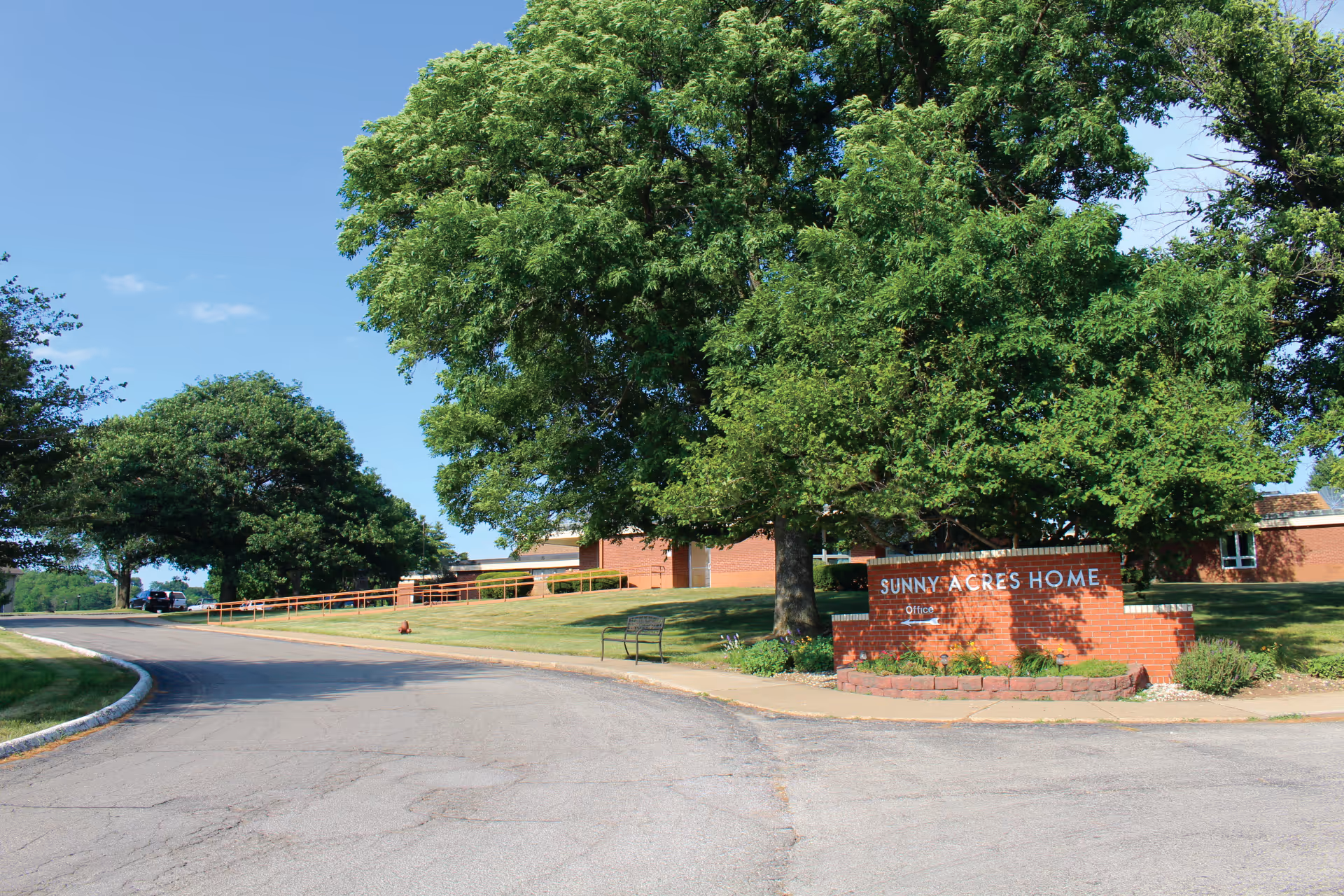 Exterior view of Sunny Acres Nursing Home showing a brick sign with the facility name, surrounded by green trees and a well-maintained lawn under a clear blue sky.